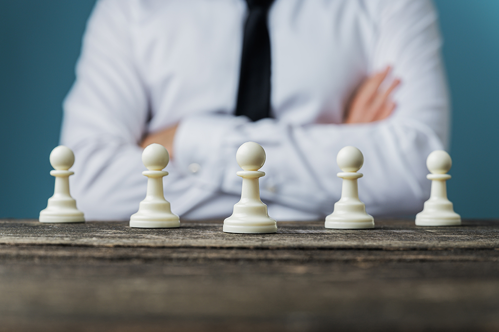 Businessman with his arms crossed sitting behind a desk with chess pawn pieces placed on it in a conceptual image.